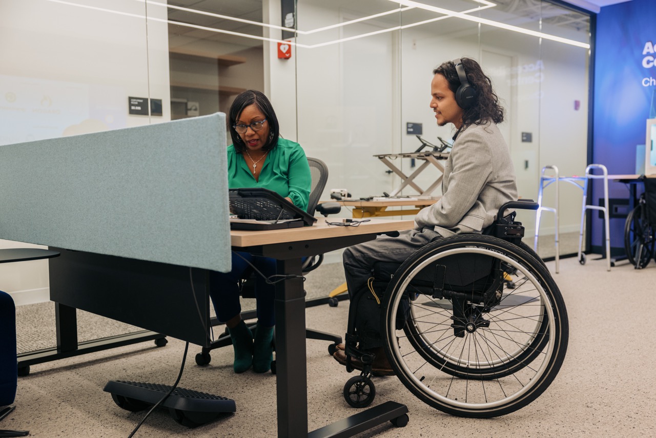 Two colleagues collaborating at a desk, one using a wheelchair and wearing headphones